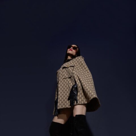 Eine Frau in stylischen schenkelhohen Stiefeln und einer gemusterten Jacke posiert dramatisch vor einem dunkelblauen Hintergrund.