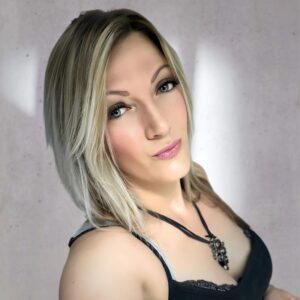 Eine Person mit glattem, schulterlangem blondem Haar trägt ein schwarzes ärmelloses Oberteil und eine Halskette und posiert für ein Selfie vor einem hellen Hintergrund.