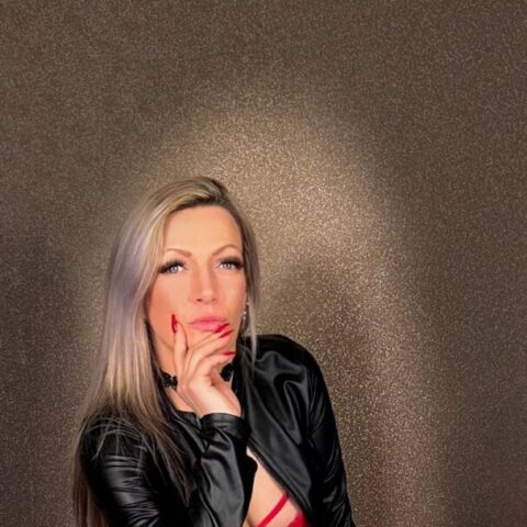 Eine Person mit langen blonden Haaren posiert in einem Innenraum, trägt eine schwarze Jacke, ein rotes Outfit und rote Overknee-Stiefel vor einer braunen, strukturierten Wand.