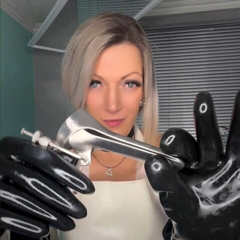 Eine Frau mit weißer Schürze und schwarzen Handschuhen hält ein medizinisches Spekulum aus Metall vor ein Fenster mit geschlossenen Jalousien.