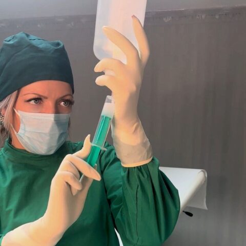 Eine medizinische Fachkraft in Kittel und Handschuhen bereitet eine Infusion vor, indem sie eine Spritze mit einer grünen Flüssigkeit in einer Klinik füllt.