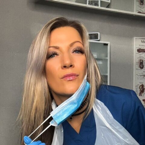 Eine Frau in medizinischem Kittel, Handschuhen und einer Plastikschürze zieht sich eine blaue Gesichtsmaske vom Gesicht und steht in einem klinischen Umfeld.