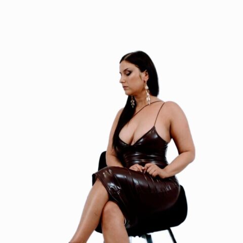 Eine Frau in einem dunkelbraunen, ärmellosen, taillierten Kleid sitzt mit übereinandergeschlagenen Beinen auf einem schwarzen Stuhl, trägt dazu passende High Heels und baumelnde Ohrringe vor einem schlichten weißen Hintergrund.