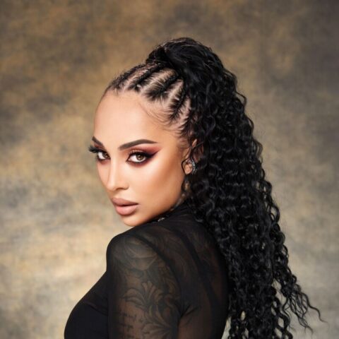 Frau mit langen, gelockten schwarzen Haaren, die zu Cornrows und einem hohen Pferdeschwanz frisiert sind. Sie trägt ein durchsichtiges schwarzes Oberteil und einen strukturierten Gürtel mit hoher Taille und steht vor einem neutralen gesprenkelten Hintergrund.