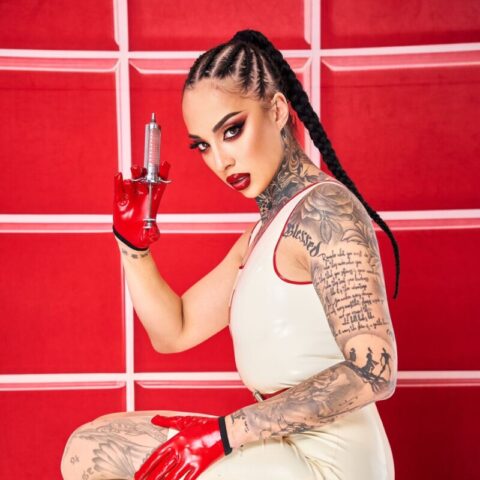 Eine tätowierte Frau mit geflochtenem Haar, die ein weißes Latexkleid, rote Handschuhe und rote High Heels trägt, hockt mit einer Spritze in der Hand vor einem roten Hintergrund.