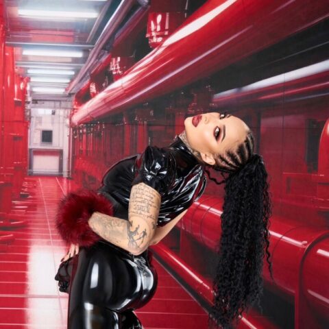Person in schwarzem Latex-Outfit und hohen Plateaustiefeln posiert in einem roten, futuristischen Korridor, mit langen geflochtenen Haaren und sichtbaren Tätowierungen an den Armen.