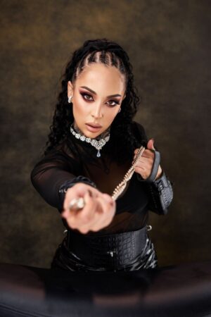 Frau mit geflochtenem Haar, auffälligem Make-up und schwarzem Outfit hält eine Leine in Richtung Kamera vor einem dunklen, strukturierten Hintergrund.