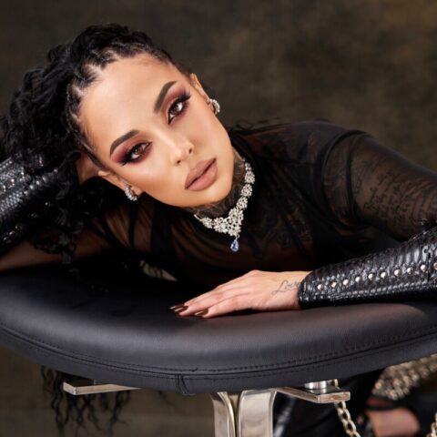 Eine Frau mit geflochtenen Haaren und dramatischem Make-up lehnt auf einem schwarzen, gepolsterten Stuhl, trägt ein durchsichtiges schwarzes Oberteil, eine juwelenbesetzte Halskette und metallische Accessoires und blickt direkt in die Kamera.