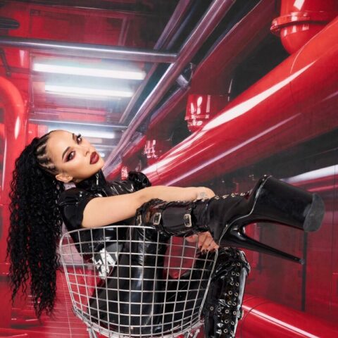 Eine Person mit langen lockigen Haaren lehnt sich in einem Metalldrahtstuhl zurück, trägt schwarze Latexkleidung und hohe Plateaustiefel vor einem roten Hintergrund im Industriestil.