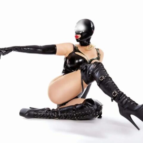 Person in schwarzem Latex-Outfit, schenkelhohen Stiefeln und Vollgesichtsmaske mit rotem Ballknebel, kniet und zeigt mit ausgestrecktem linken Arm.