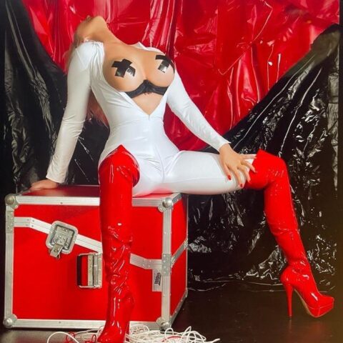 Eine Person in einem weißen Outfit mit schwarzem Klebeband über der Brustpartie und roten, schenkelhohen Stiefeln posiert auf einem roten und metallischen Koffer. Schwarz und rot drapierter Hintergrund.