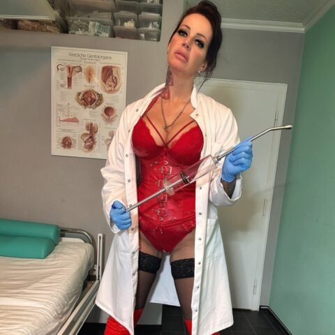 Eine Person in roter Unterwäsche, Strümpfen und kniehohen Stiefeln trägt einen weißen Laborkittel und blaue Handschuhe und steht in einem Behandlungszimmer und hält ein Metallinstrument in der Hand.