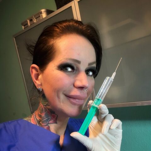 Eine Person in blauer Arztuniform, mit Tätowierungen am Hals, hält eine mit grüner Flüssigkeit gefüllte Spritze, trägt weiße Handschuhe und lächelt in die Kamera.