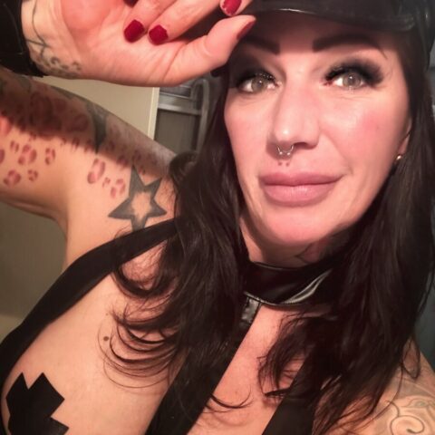 Eine Person mit langen Haaren und Tattoos, die ein schwarzes Outfit, einen Hut und einen Nasenring trägt, posiert in einem Innenbereich für ein Selfie.