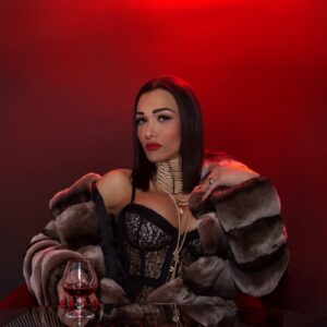 Eine Frau in Spitzentop und Pelzmantel sitzt mit einem Glas Rotwein vor einem roten Hintergrund an einem Tisch.