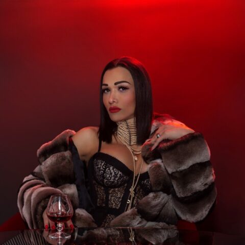 Eine Frau in Spitzentop und Pelzmantel sitzt mit einem Glas Rotwein vor einem roten Hintergrund an einem Tisch.