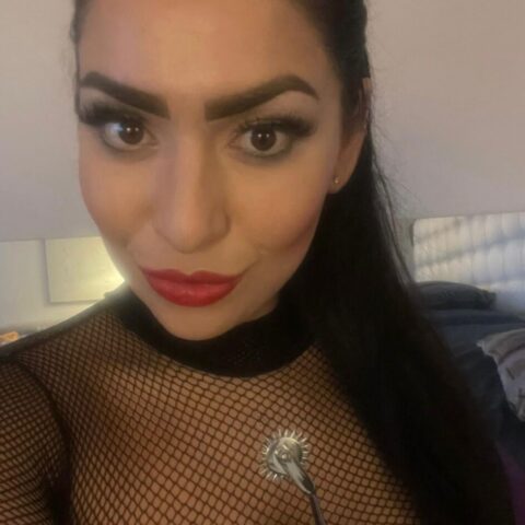 Eine Frau, die ein schwarzes Netzoberteil und Handschuhe trägt, hält sich ein mit Stacheln besetztes Metallwerkzeug an die Brust und blickt mit auffälligem Make-up in die Kamera.