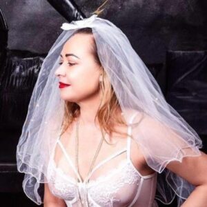 Person, die in einem schwach beleuchteten Raum auf einem Kissen kniet und ein weißes Spitzenoutfit mit Schleier und Strumpfband trägt.