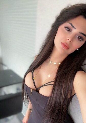 Eine Frau mit langen dunklen Haaren und einem schwarzen Kleid steht in einem Haus und schaut in die Kamera. Im Hintergrund sind ein verschwommener Tisch und Stühle zu sehen. Ortsangabe: München, Deutschland.