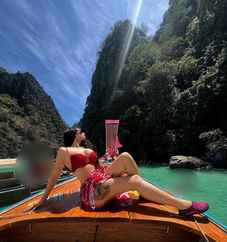 Eine Frau im roten Bikini sitzt am Bug eines Holzbootes in türkisfarbenem Wasser, umgeben von steilen, grünen Klippen unter blauem Himmel.