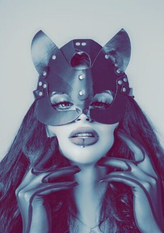 Person, die eine lederne Katzenmaske, lange Handschuhe und dramatisches Make-up trägt und mit den Händen in der Nähe des Gesichts posiert; das Bild hat einen bläulichen Farbton.
